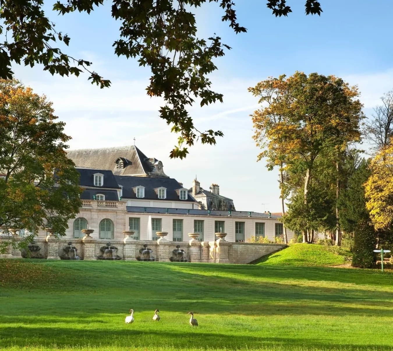 Exterior of the Auberge du Jeu de Paume in Chantilly, a stone château facade above a lawn with swans on a sunny day