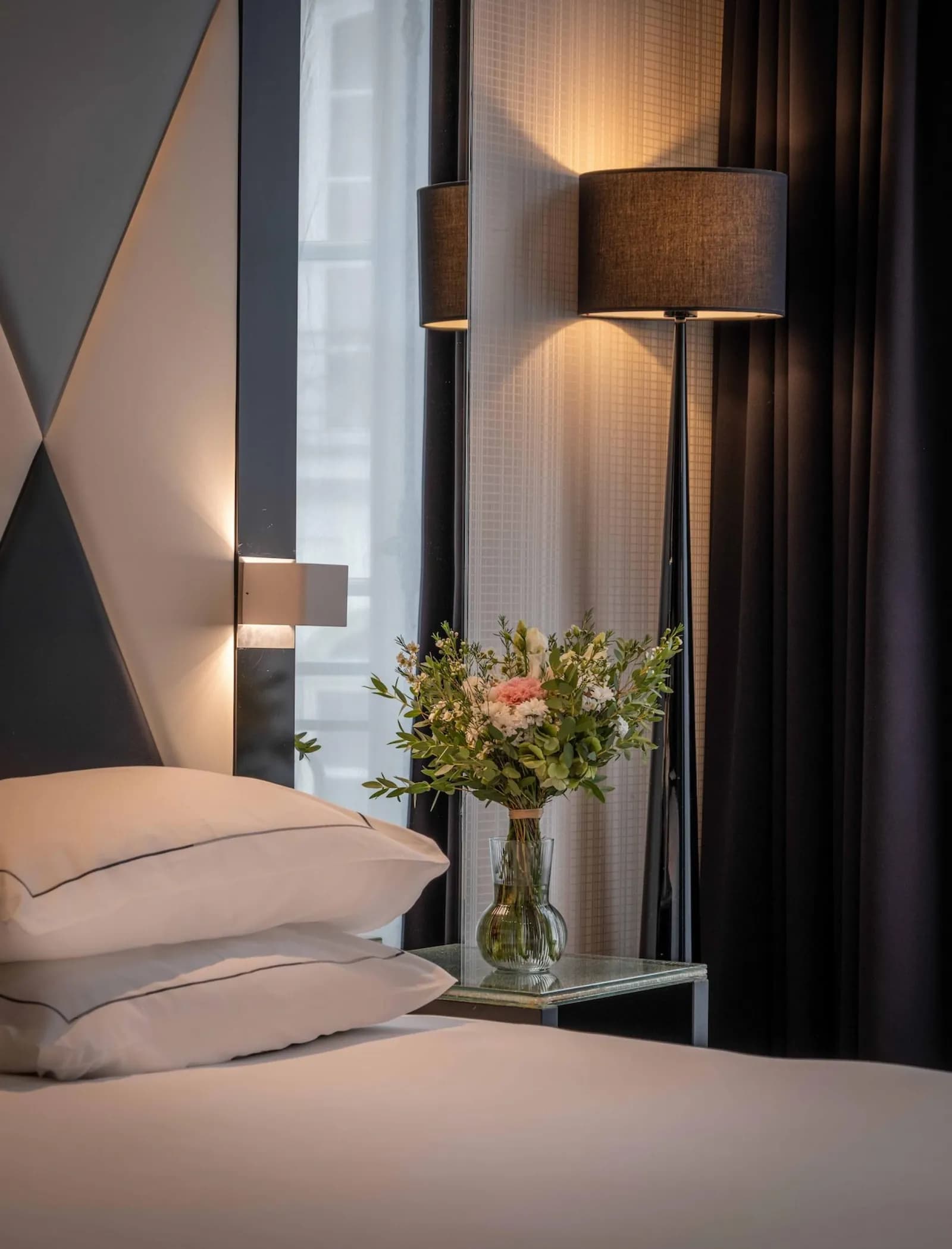 Bedroom with a grey geometric headboard, white pillows and a flower vase beside a lamp at L'Empire Paris