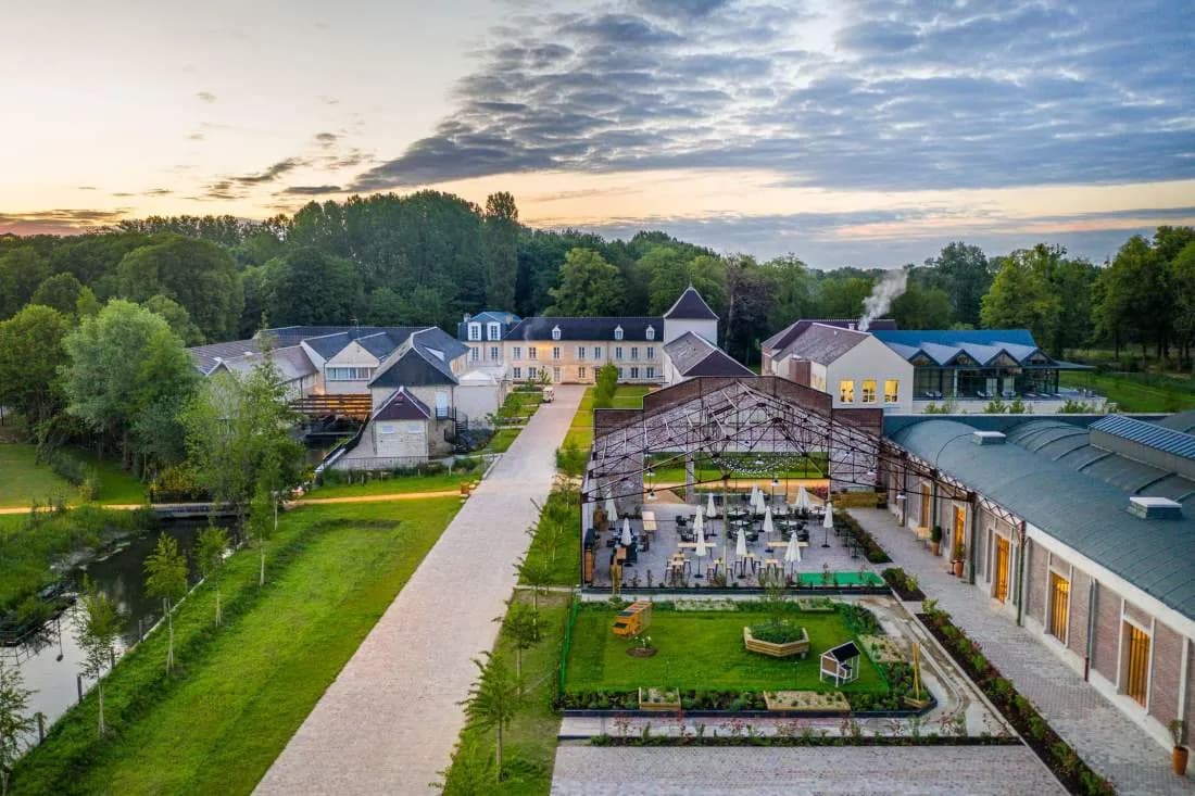 Aerial view of Le Grand Pavillon Chantilly, a stone estate with formal gardens, a glass-roofed terrace and a river