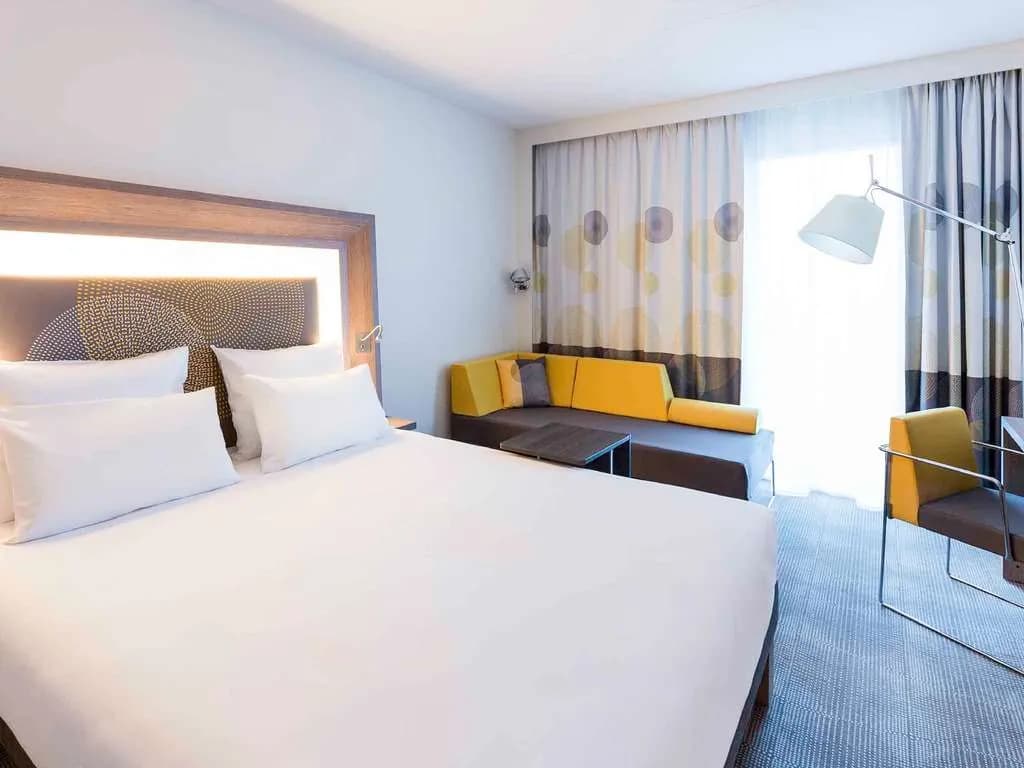 Double room with yellow accent armchair, sofa bed and wide window at Novotel Bordeaux Lac