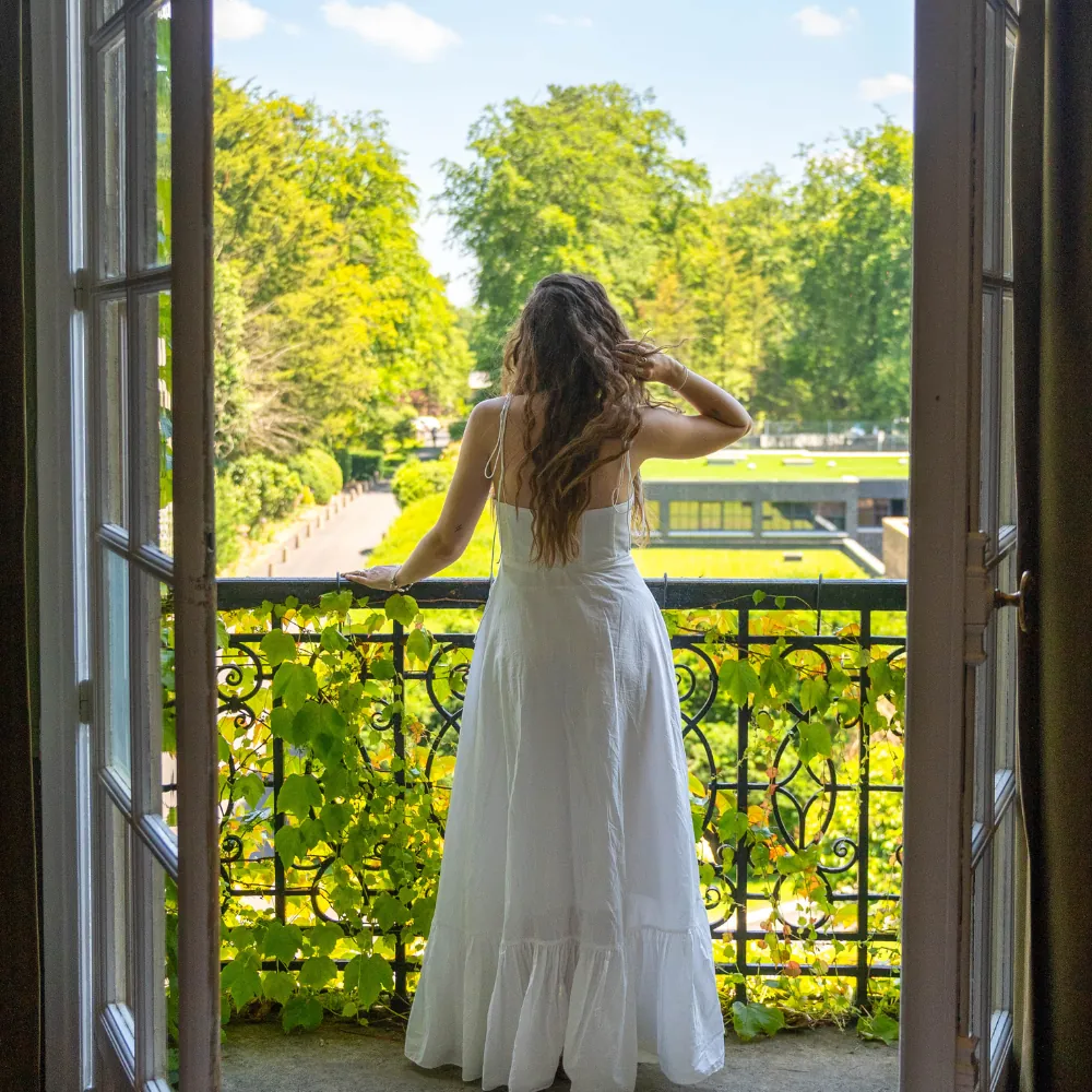 Guest on a wrought-iron balcony of Château Jeanne & The Forest, looking over the green park and trees of the estate