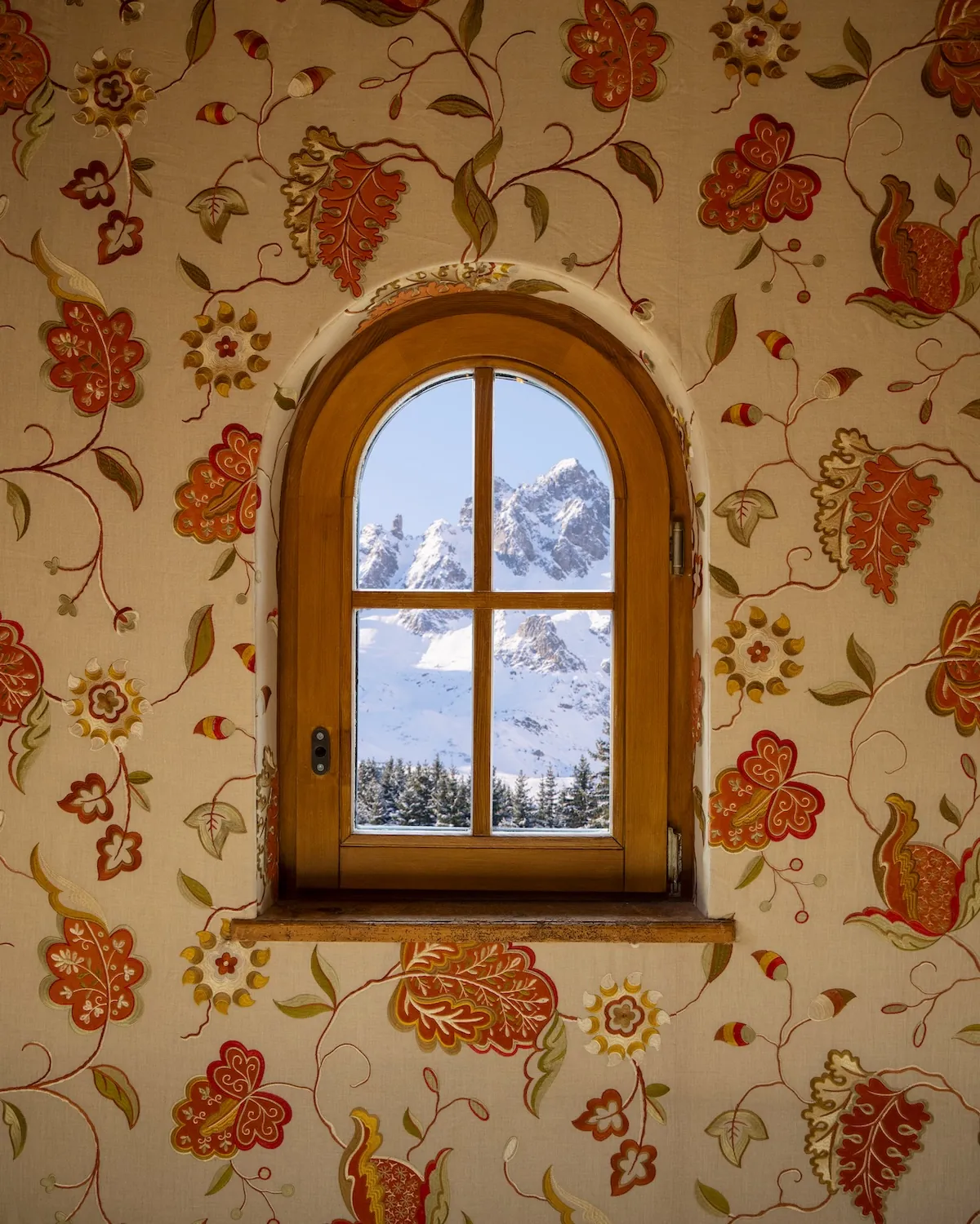 Arched window framing snowy peaks, with floral wallpaper, at Les Airelles Courchevel, Courchevel 1850