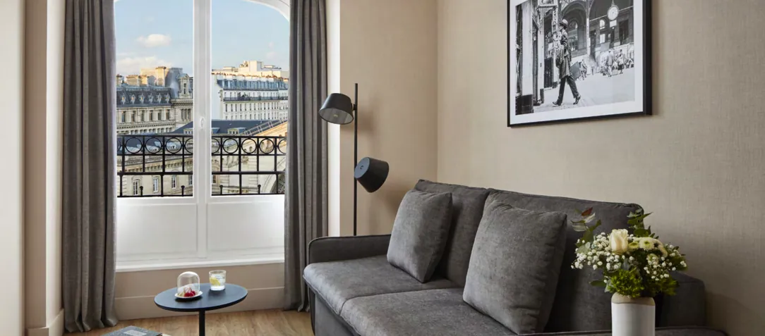 Guest room at the NH Paris Gare de l'Est with a grey sofa and a window overlooking the Paris rooftops and balconies