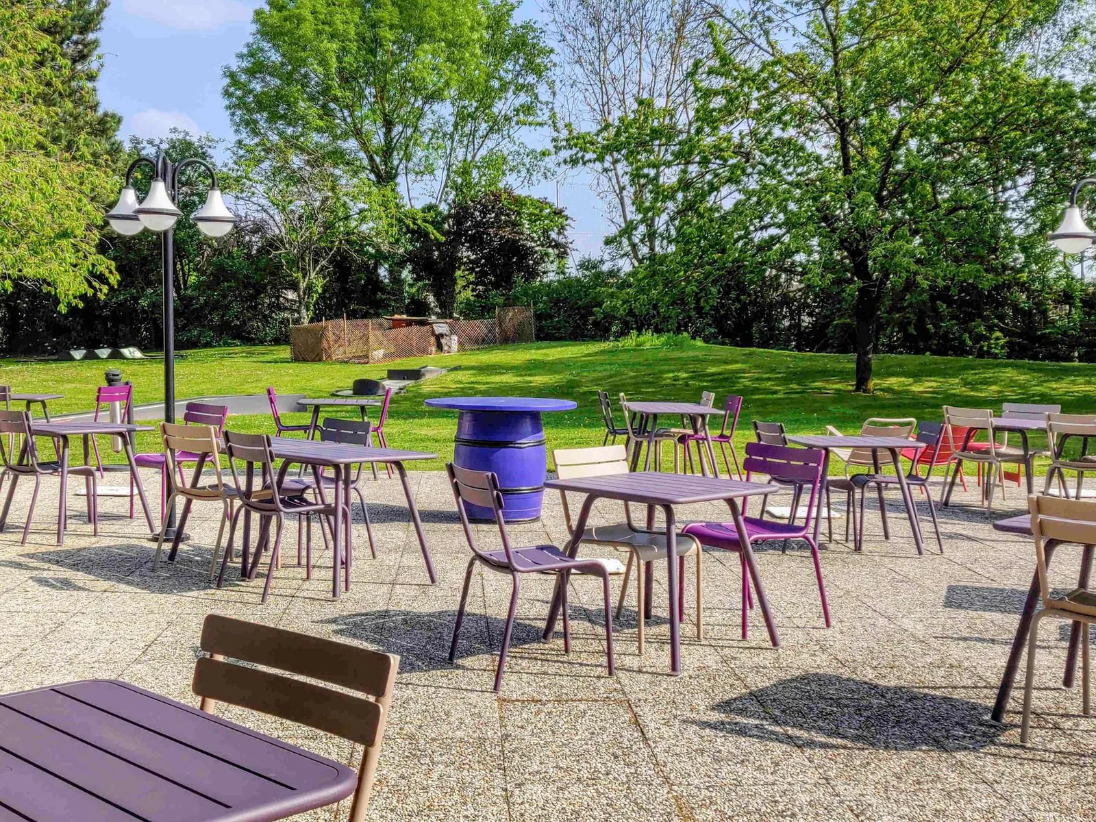 Shaded terrace with purple tables, a green lawn and tall trees at Novotel Saclay, Essonne