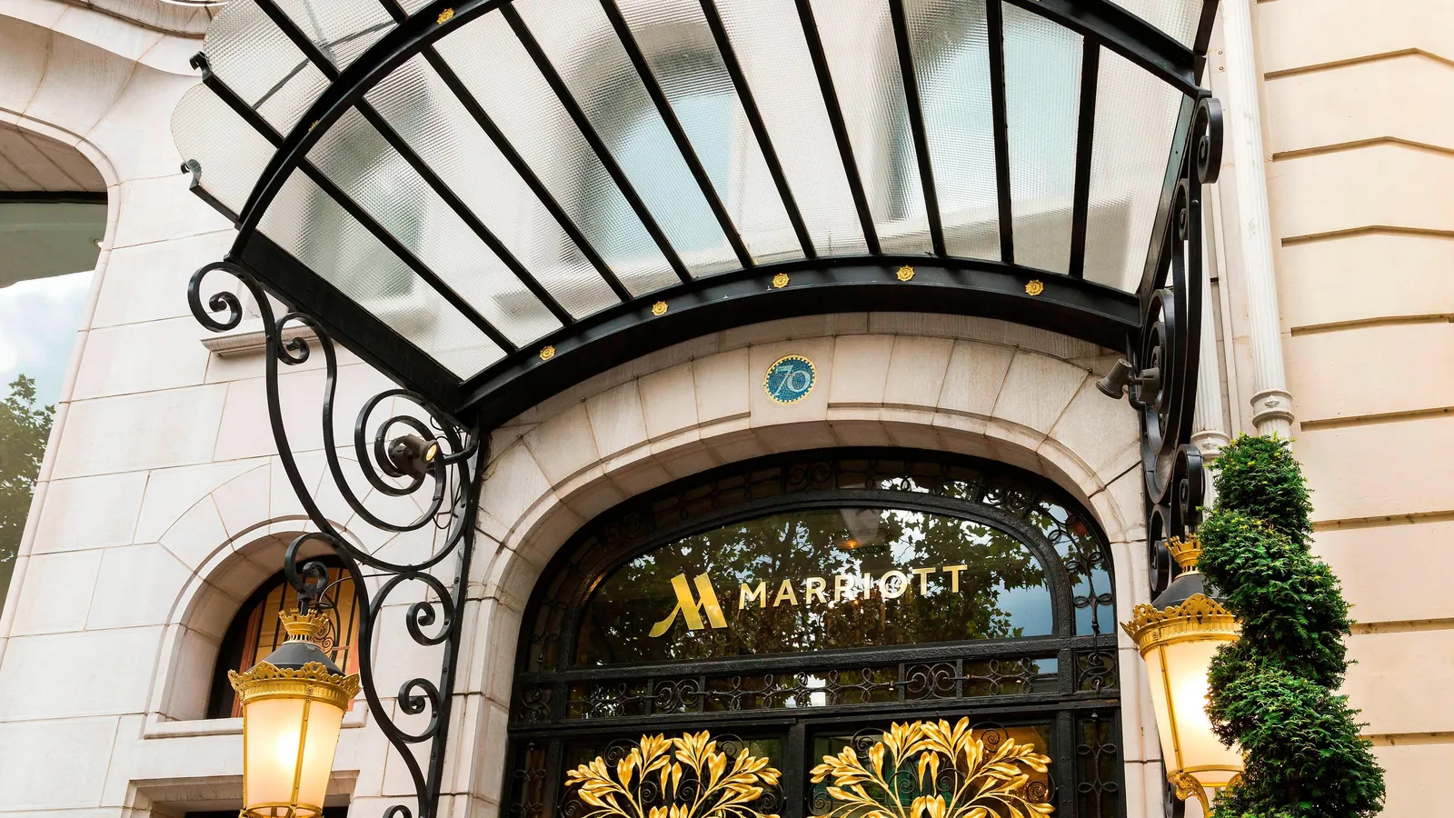 Glass canopy and golden Marriott entrance doors of the Paris Marriott Champs-Elysees Hotel, framed by stone and topiary