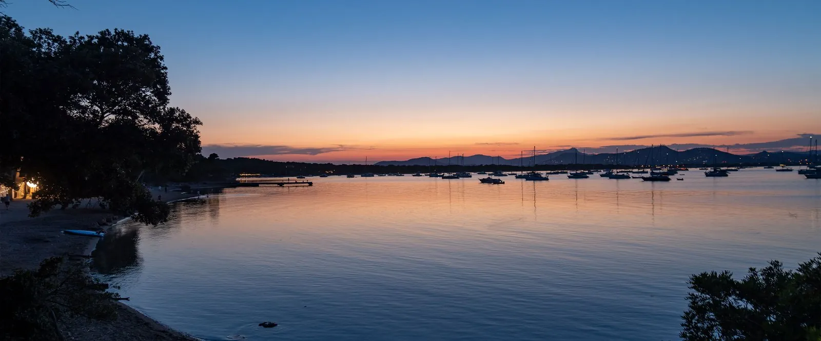 Sunset over a calm bay with moored boats and distant hills, seen from Résidence des Pins Méditerranéens, Var