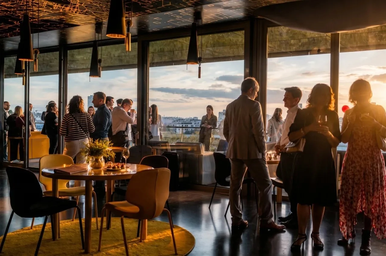 Guests at a cocktail reception beside tall windows at sunset