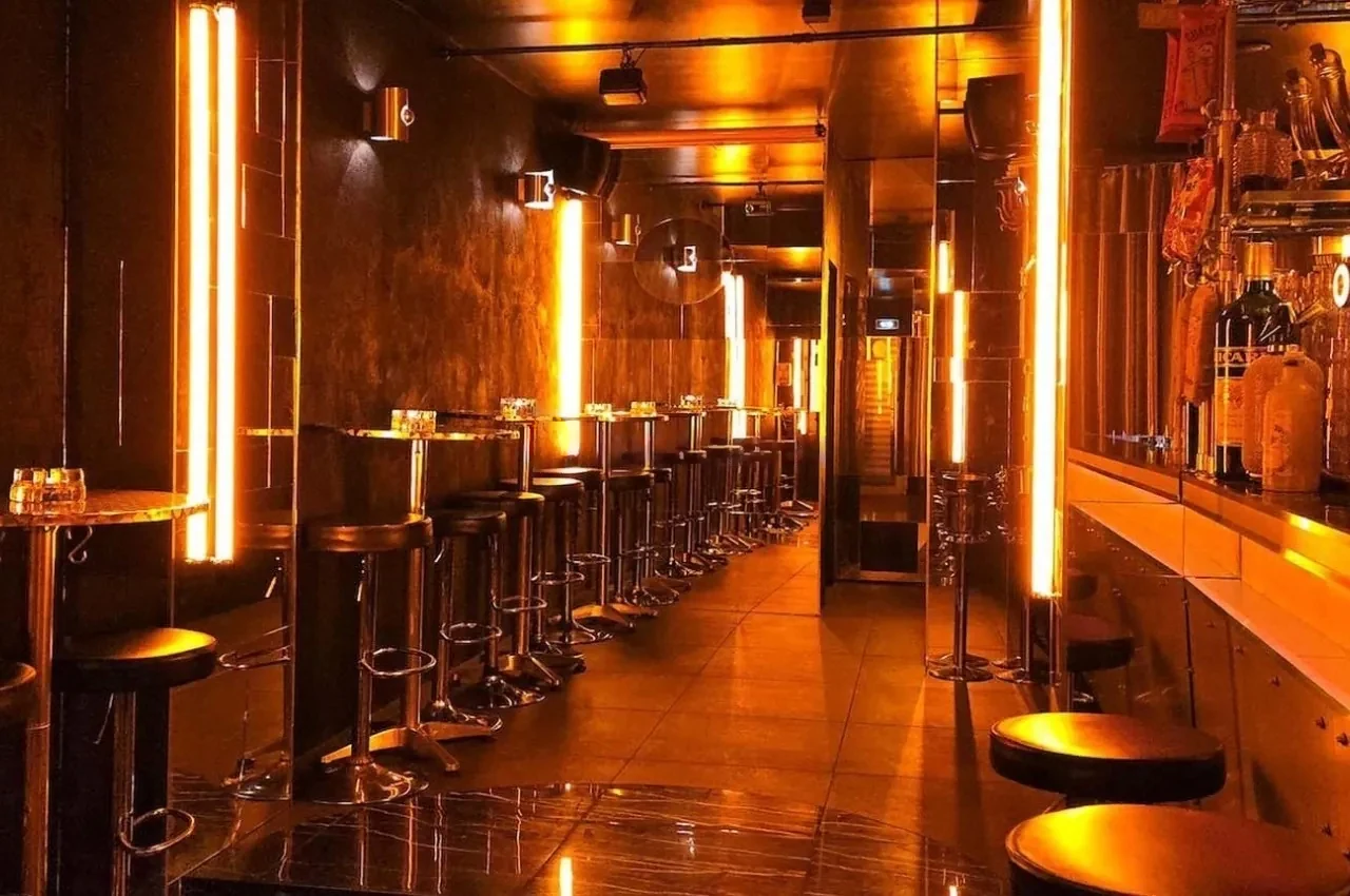 Bar lit by amber neon tubes with mirrored shelves and a counter of bottles.