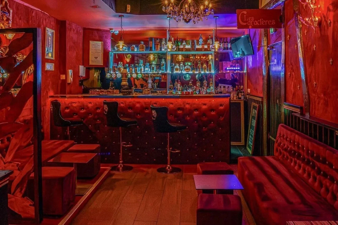 Bar with red velvet banquettes, a chandelier and illuminated bottle shelves.