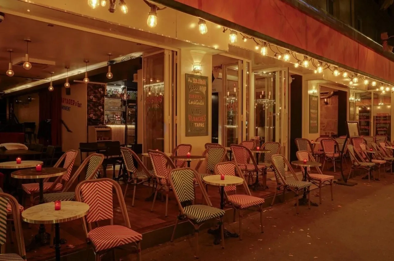 Street terrace of a venue at dusk with striped chairs, string lights and a chalkboard menu.